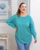 Woman wearing a teal sweater and blue jeans standing in a room with a decorative wall sign.