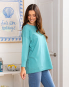 Woman wearing a teal long-sleeve top and blue jeans in a bathroom.