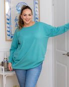 Woman wearing a teal sweater and blue jeans standing in a room with a decorative wall.