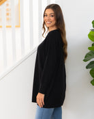 Woman wearing a black sweater and blue jeans standing against a white wall with a plant to the side.