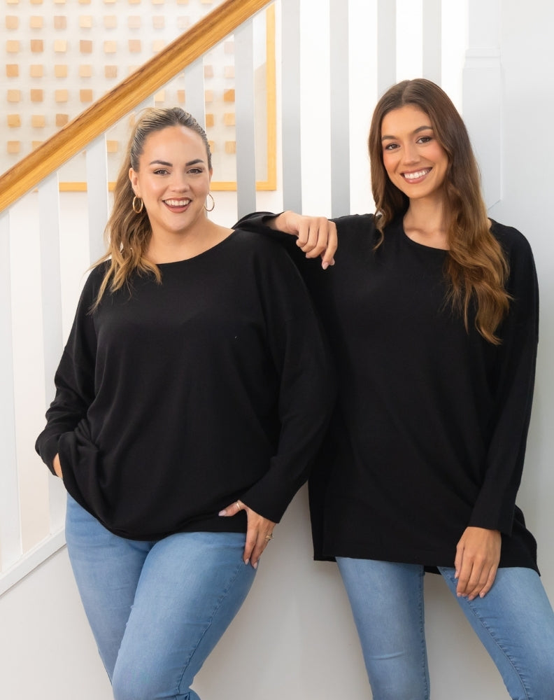 Two women wearing black oversized tunic knit and blue jeans standing against a white wall.