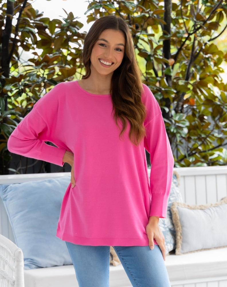 Woman wearing a bright pink long-sleeve top standing outdoors with greenery in the background.
