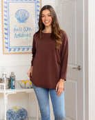 Woman wearing a brown long-sleeve top and blue jeans standing in a room with a decorative wall sign.