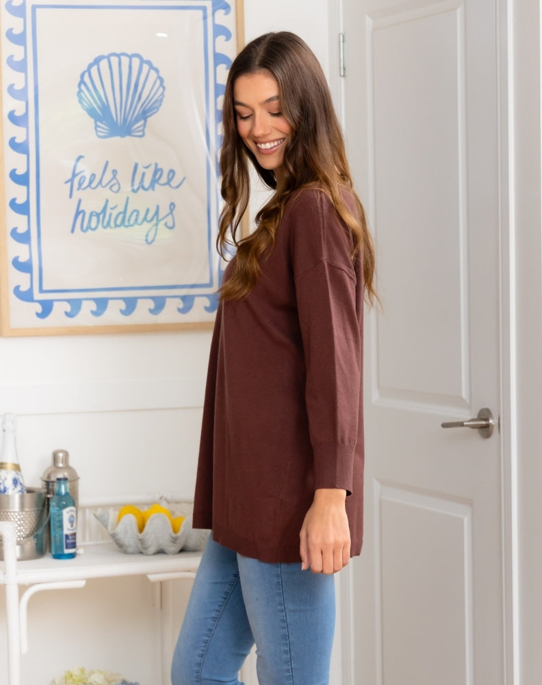 Woman standing in a kitchen wearing a brown cardigan and blue jeans, with a decorative sign on the wall.