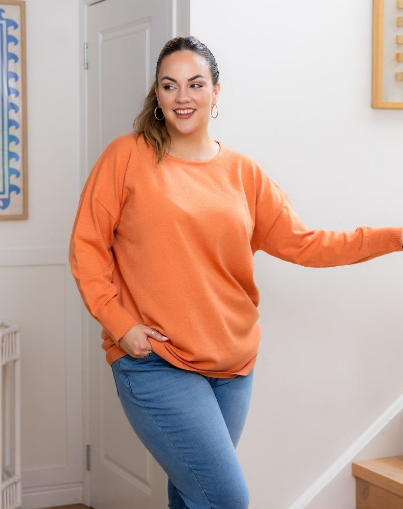 Woman wearing an orange sweater and blue jeans standing indoors.