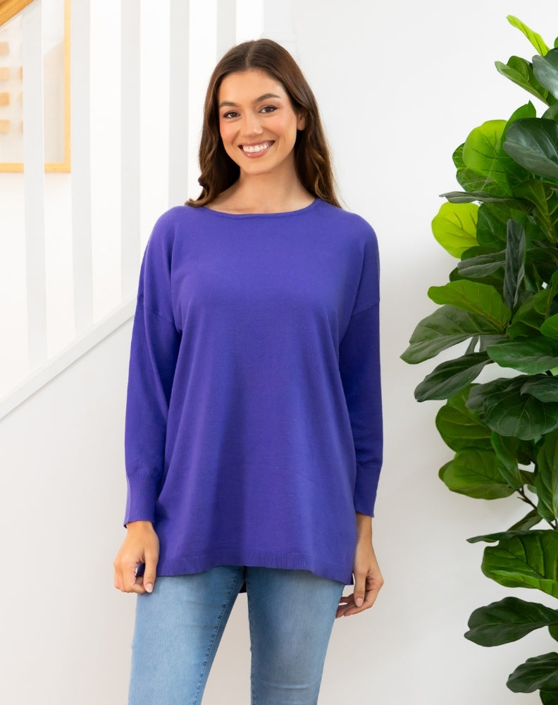 Woman wearing a purple long-sleeve top standing next to a green plant indoors.