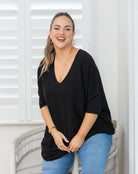 A woman smiling while wearing a black, loose-fitting V-neck top with three-quarter sleeves and light blue jeans, sitting on a white wicker chair in front of white shuttered windows.