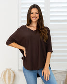A woman wearing a dark brown, loose-fitting V-neck top with three-quarter sleeves and blue jeans, standing in front of white window blinds.