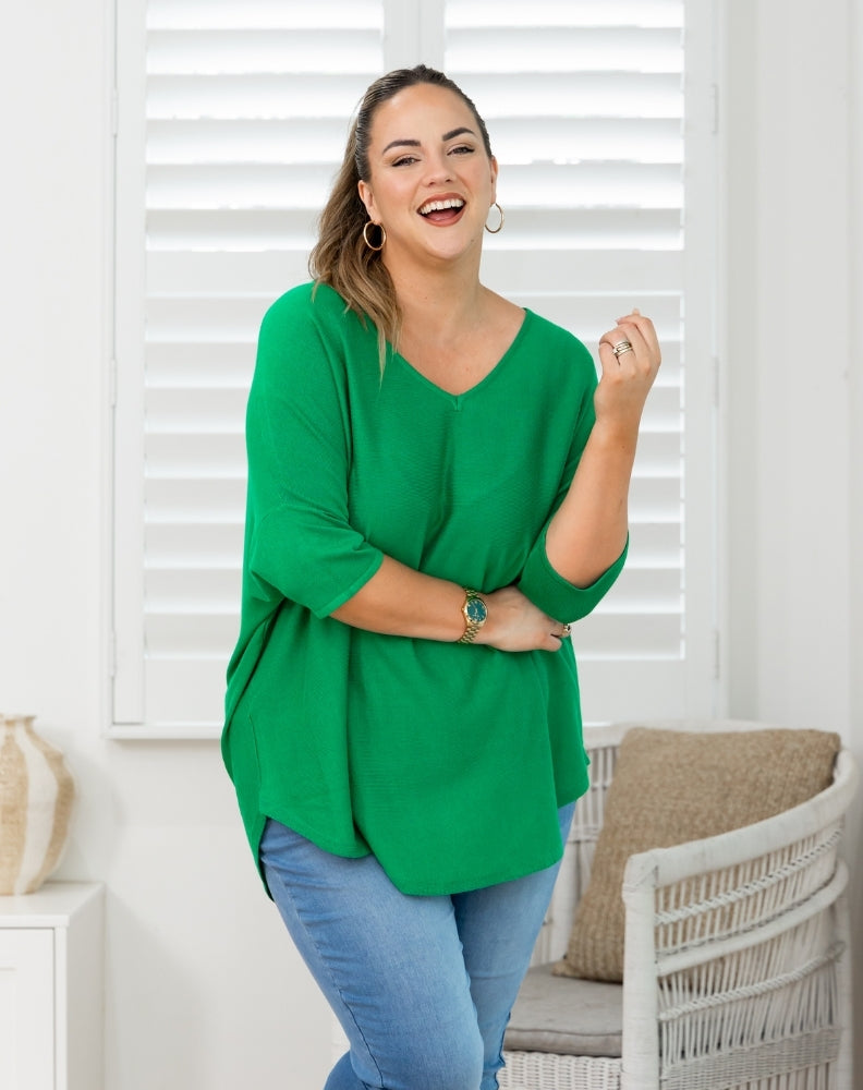A woman modeling a bright green three-quarter sleeve V-neck top with blue jeans, standing in a brightly lit room with white shutters and a wicker chair.