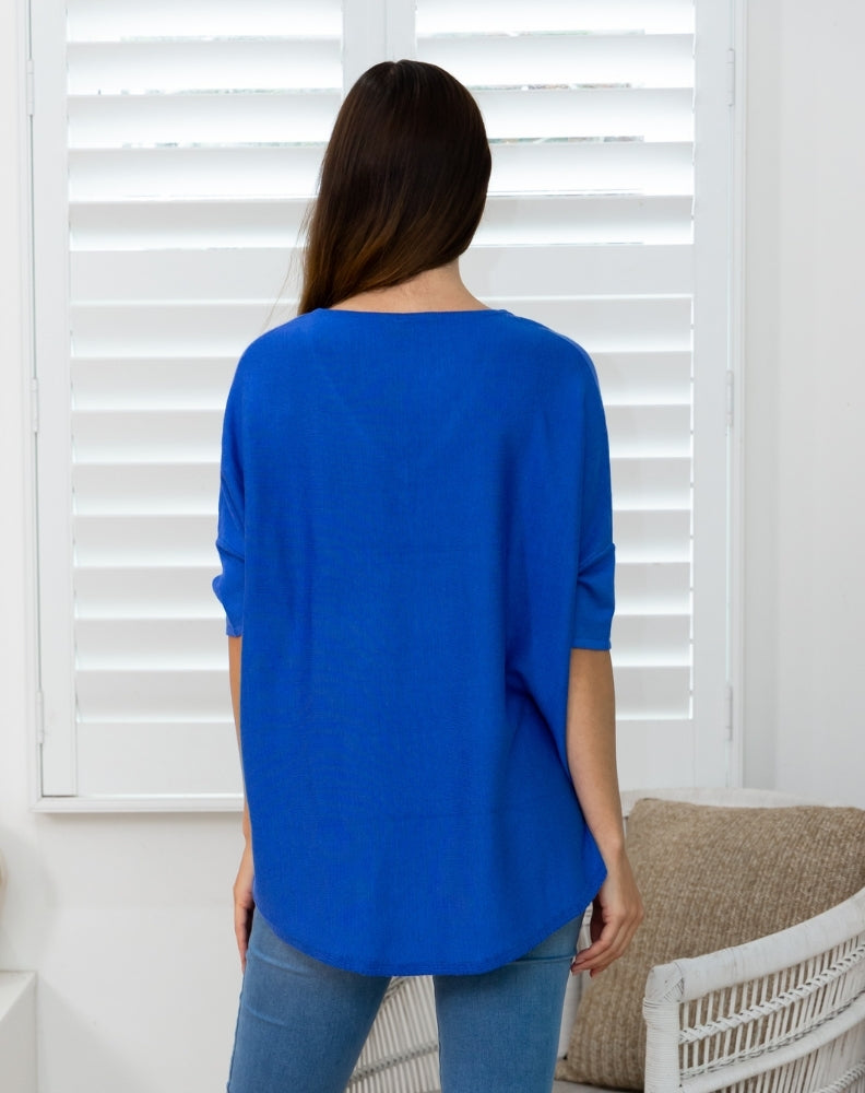 A woman viewed from behind wearing a bright royal blue, three-quarter sleeve top and light blue jeans, standing in front of white window shutters.