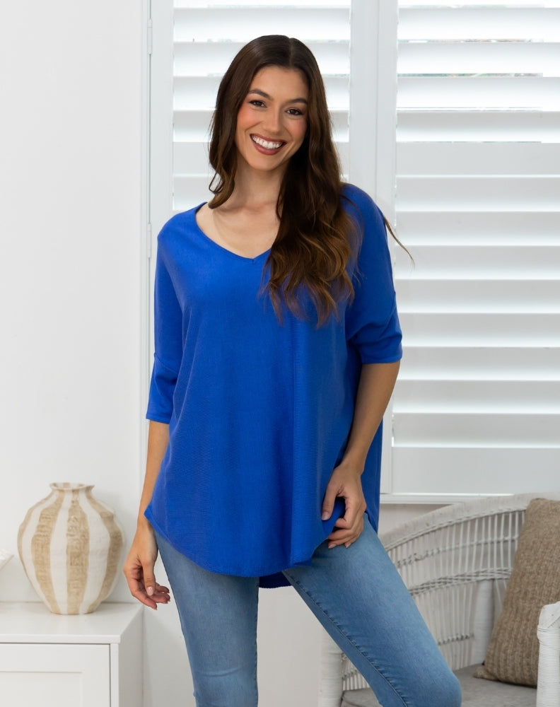 A woman wearing a bright royal blue three-quarter sleeve top and light wash blue jeans, standing in a brightly lit room with white window shutters.
