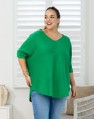 A woman modeling a bright green V-neck top with three-quarter sleeves and a side slit, paired with blue jeans, standing in front of white window shutters.