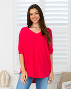 A woman wearing a bright red V-neck tunic top with three-quarter sleeves and blue jeans, standing in front of white window shutters.