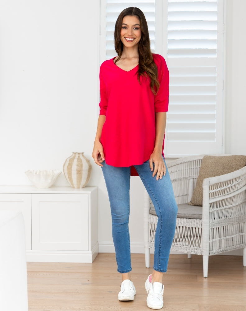 A woman modeling a bright red, three-quarter sleeve V-neck tunic top paired with blue skinny jeans and white sneakers, standing in a bright, modern room with white shutters and a wicker chair.