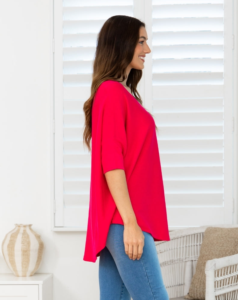 A woman wearing a bright pinkish-red short-sleeve V-neck top and blue jeans, standing in profile in front of white window shutters.