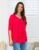 A woman wearing a bright red V-neck top with three-quarter sleeves and blue jeans, standing in front of white window shutters.