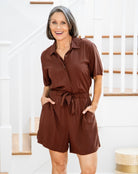 Woman wearing a brown romper standing in a home setting with a staircase in the background.