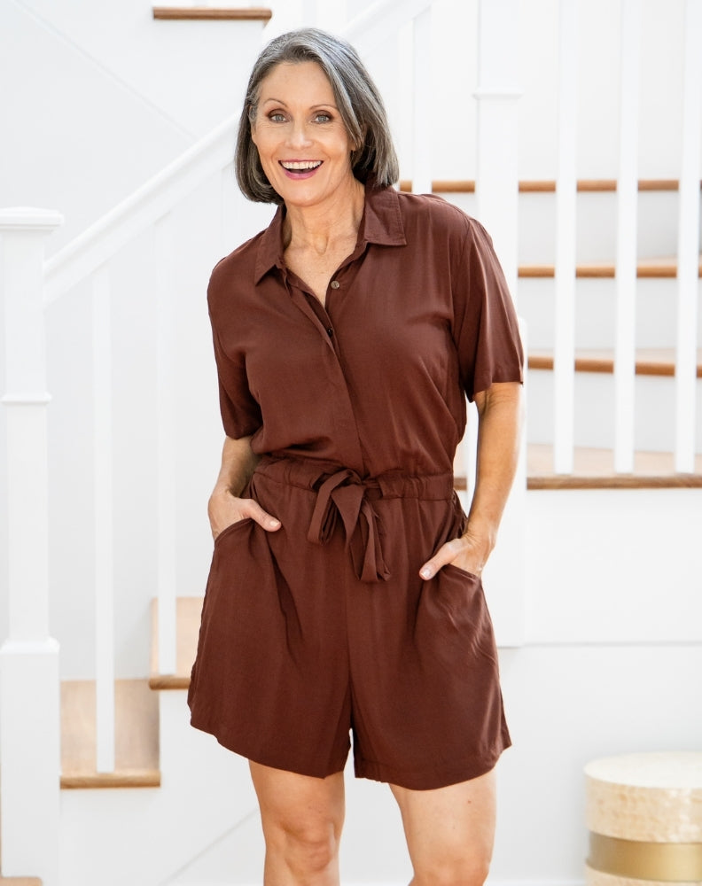 Woman wearing a brown romper standing in a home setting with a staircase in the background.