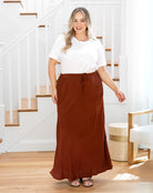 Woman wearing a white top and brown maxi skirt standing in a home setting.
