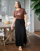 Woman standing on a wooden deck wearing a brown t-shirt with a graphic design and a black skirt.