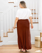Person wearing a white top and brown skirt standing in a home setting with stairs in the background.