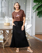 Woman wearing a brown t-shirt and black skirt standing on a wooden deck.