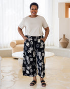 Woman wearing a white top and black patterned pants in a room with a couch and vase.