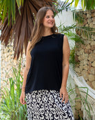 Woman wearing a black sleeveless top and patterned skirt standing outdoors with plants and a stone wall in the background.