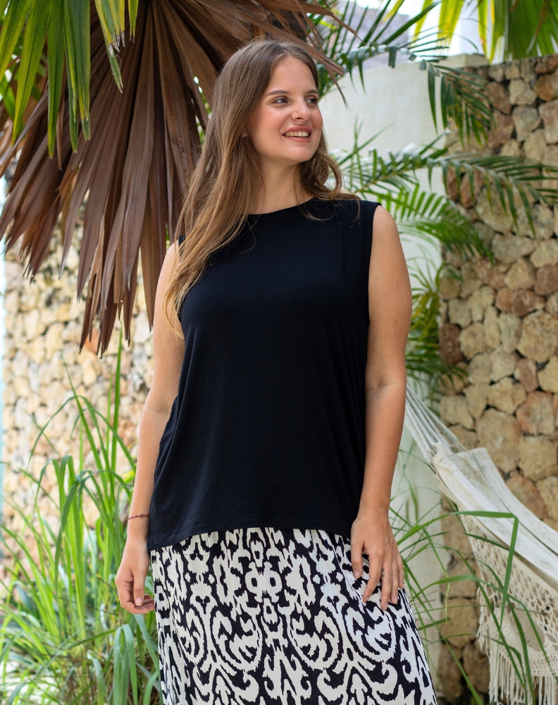 Woman wearing a black sleeveless top and patterned skirt standing outdoors with plants and a stone wall in the background.