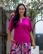 Woman wearing a bright pink top and floral skirt standing outdoors with greenery in the background