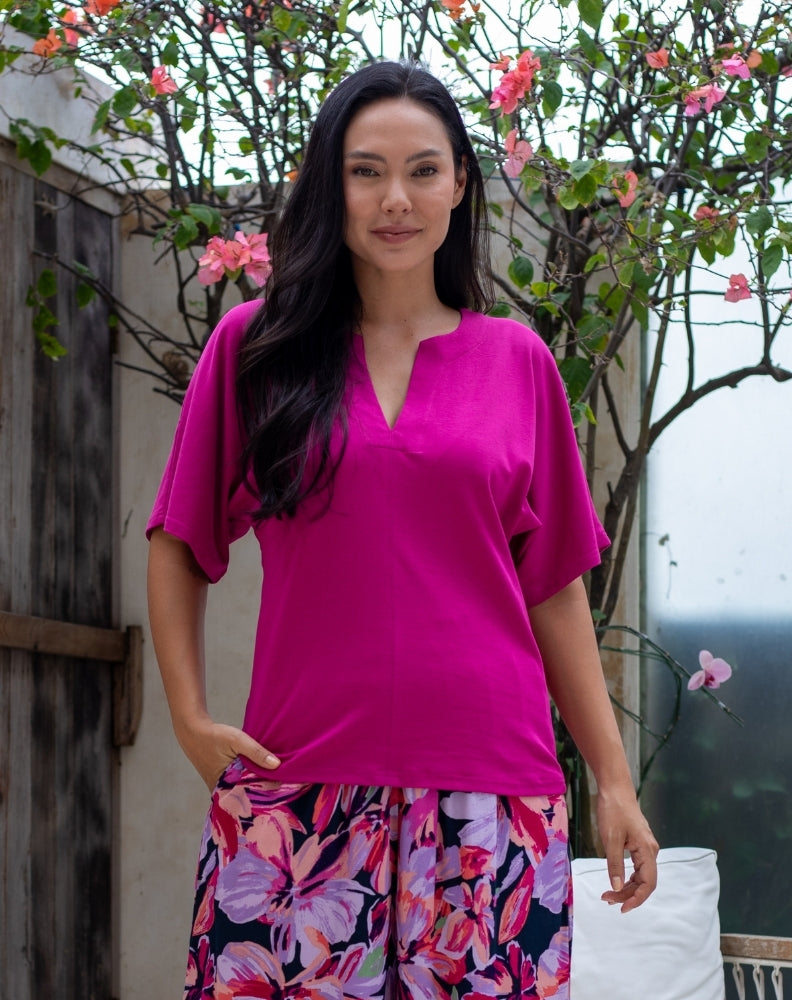 Woman wearing a bright pink top and floral skirt standing outdoors with greenery in the background