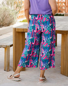 A person wearing colorful wide-leg cropped pants with a bold geometric pattern in pink, teal, blue, and white, paired with a purple top and beige sandals, standing outdoors near wooden tables.