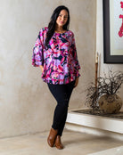 Woman wearing a colorful floral blouse and black pants standing indoors.
