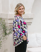 A woman wearing a black long-sleeve blouse with a vibrant multicolor floral pattern of pink, purple, white, and yellow flowers, paired with black trousers, standing in front of a white stone column.