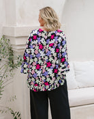 A woman shown from the back wearing a black blouse with a colorful floral pattern in pink, purple, and white, paired with black wide-leg pants, standing in a bright, modern interior setting.