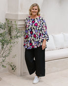 A woman modeling a black blouse with a vibrant multicolor floral pattern in pink, purple, and white, paired with black wide-leg pants and white sneakers, standing in a bright, modern indoor setting with a white sofa and stone columns.