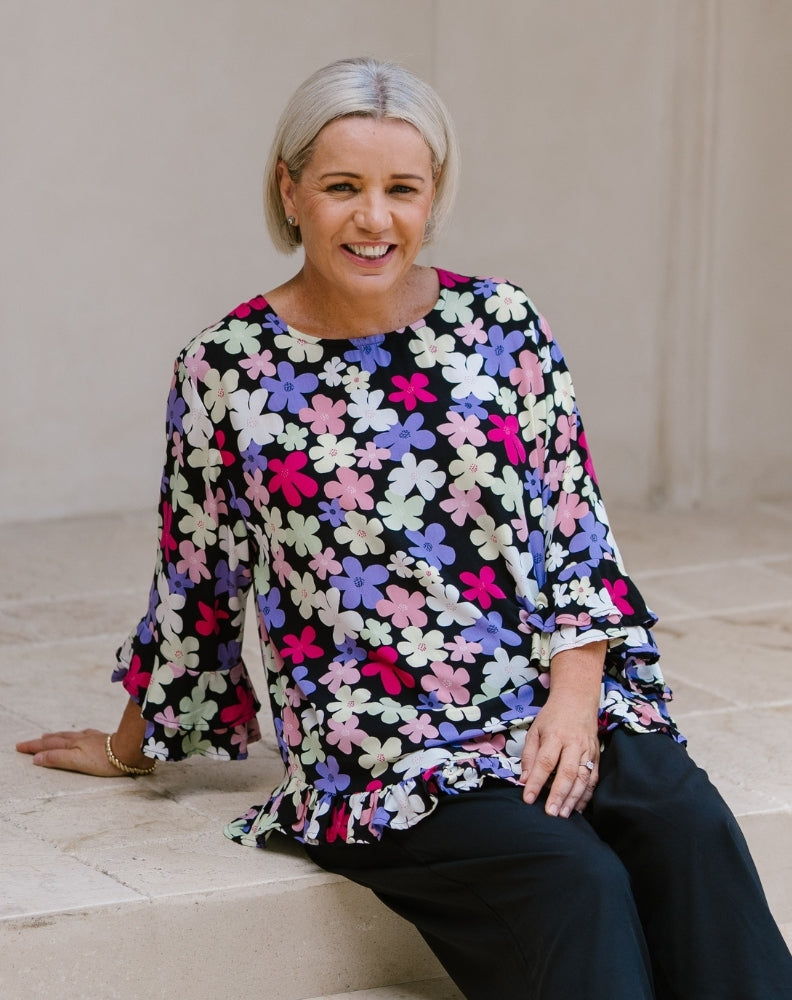 A woman with short gray hair sits on stone steps wearing a black blouse with a colorful floral pattern in pink, purple, white, and yellow, paired with black pants.
