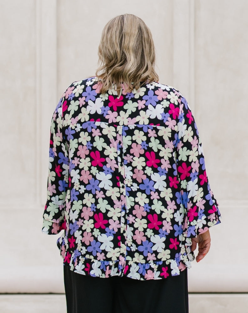 A woman viewed from behind wearing a black blouse with a vibrant multicolor floral pattern in pink, purple, and white, paired with black trousers, set against a neutral cream-colored wall.
