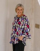 A smiling woman with short blonde hair wearing a black long-sleeve blouse with a colorful floral pattern of pink, purple, and white flowers, paired with black trousers, standing against a beige wall.