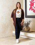 Woman wearing a brown cardigan, white shirt with a cross, and black pants in a room with a decorative vase and artwork.
