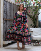 Woman wearing a colorful floral dress standing in an outdoor setting with a wooden door and white chair.