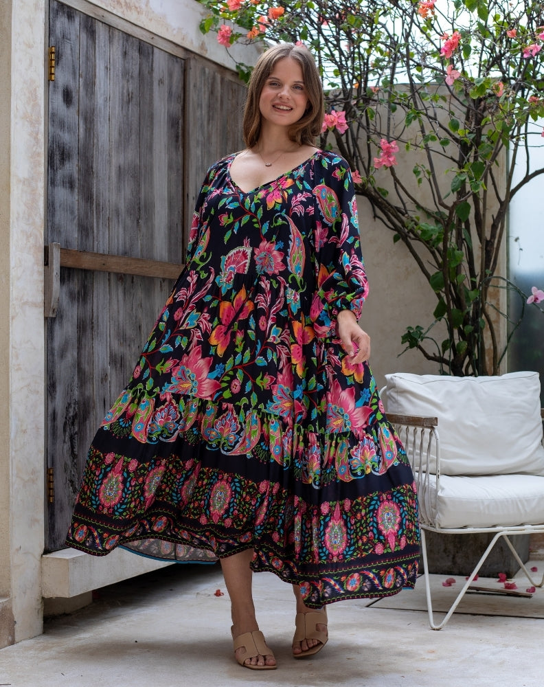 Woman wearing a colorful floral dress standing in an outdoor setting with a wooden door and white chair.