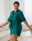 Woman wearing a teal romper in a bright room with white curtains.