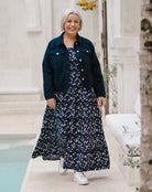 A woman with short gray hair walking outdoors in a navy blue denim jacket over a navy and white floral print maxi dress, paired with white sneakers.