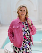 A woman wearing a bright pink denim-style jacket over a black dress with a colorful floral print, standing in front of a light-colored architectural background with a pool visible.