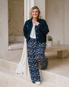 A woman posing on stone steps wearing a navy blue denim jacket, a white t-shirt, and navy blue wide-leg trousers with a white and light blue abstract pattern. She is smiling and looking directly at the camera in a bright, modern interior setting.