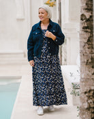 A woman with short gray hair stands outdoors wearing a navy blue denim jacket over a navy and white floral print maxi dress, paired with white sneakers. She is posing in front of a light-colored stone building with a tree trunk on the right.