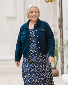 A smiling woman with short gray hair stands outdoors wearing a dark blue denim jacket over a navy and white patterned dress.