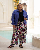 A woman modeling wide-leg floral pants, a black top, and a blue denim jacket in an outdoor patio setting with a pool in the background.