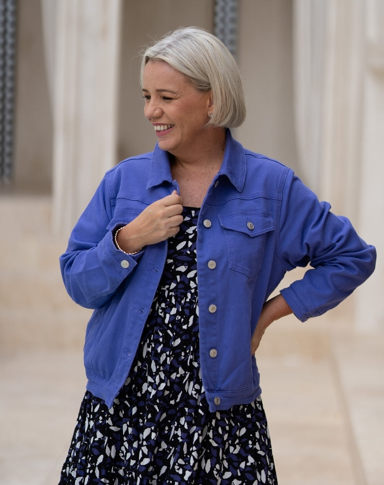 A woman with short gray hair wearing a bright blue denim jacket over a navy and white patterned dress, standing in a softly lit indoor setting with beige columns.